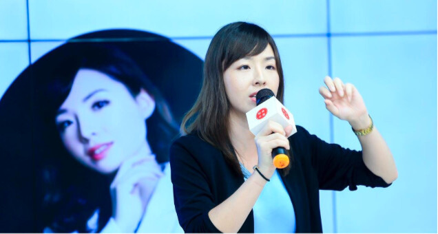 Yingying Li speaking with a microphone on stage, her portrait on the screen behind her